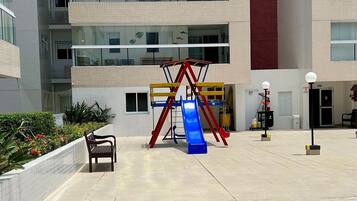 A playground with a slide and swings, surrounded by a building with multiple balconies and a tall red column.