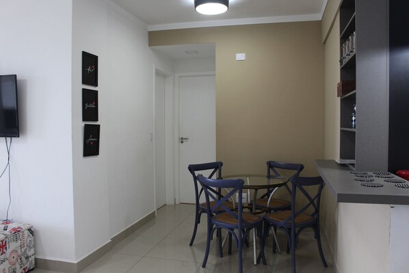 A compact dining area with a round table and four chairs, a television mounted on the wall, and a small cabinet with shelves. - Av. Anchieta 1636 (Bertioga)