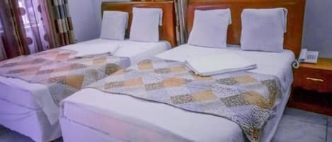 Een hotelkamer met twee bedden, elk met een gewatteerde deken en witte kussens. Er staat een nachtkastje met een telefoon en een lamp.