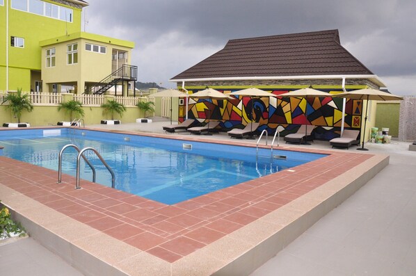 A swimming pool with lounge chairs and umbrellas, a colorful mural, and a building with a sloped roof.