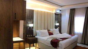 Een hotelkamer met een groot bed, bedlampen, een nachtkastje, een houten garderobe en uitzicht op het groen door een raam.