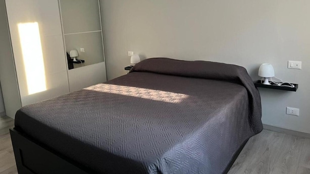 A modern bedroom with a large bed, a wardrobe, a bedside table with a lamp, and a wall-mounted shelf with a lamp. - Saint Rita's (Torino)
