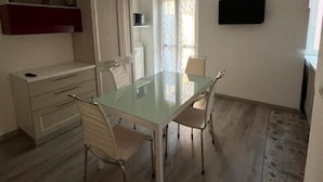 A compact living space with a glass dining table, two chairs, a TV mounted on the wall, and a kitchenette area with cabinets and a microwave. - Saint Rita's (Torino)