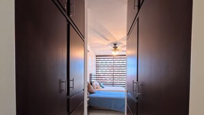 Un pasillo estrecho que lleva a un dormitorio con cama, ventilador de techo y ventana con persianas.