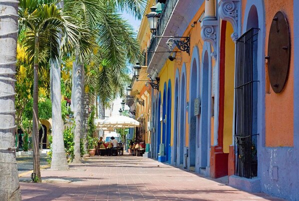 A street lined with colorful buildings, palm trees, and outdoor seating areas.