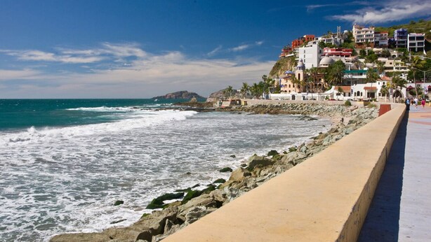 Un paseo marítimo en la costa con un litoral rocoso, un sendero de arena y edificios con balcones que dan al mar.
