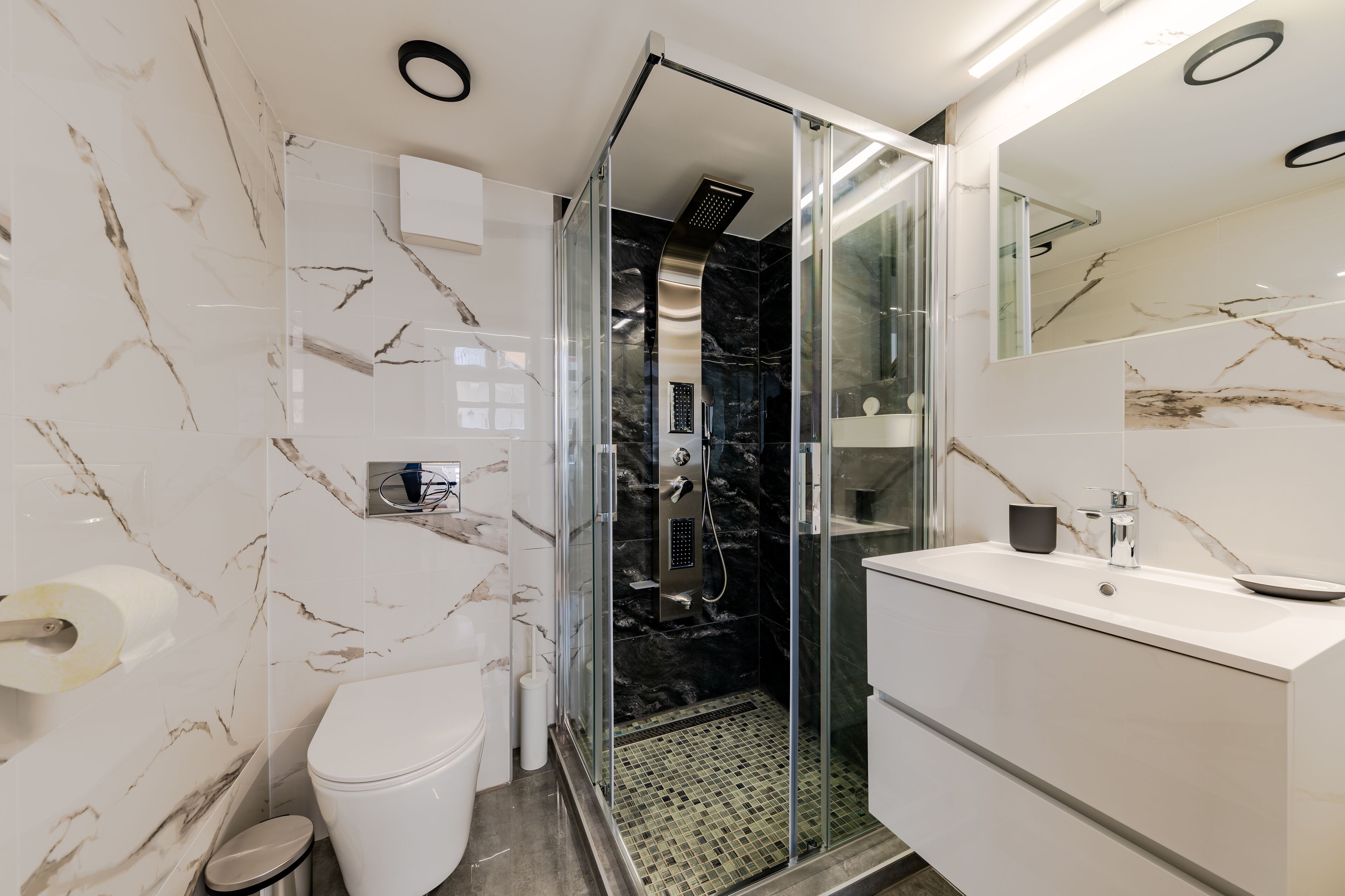 Een moderne badkamer met een glazen douchecabine, een wit wastafeltje en een marmeren spatwand.