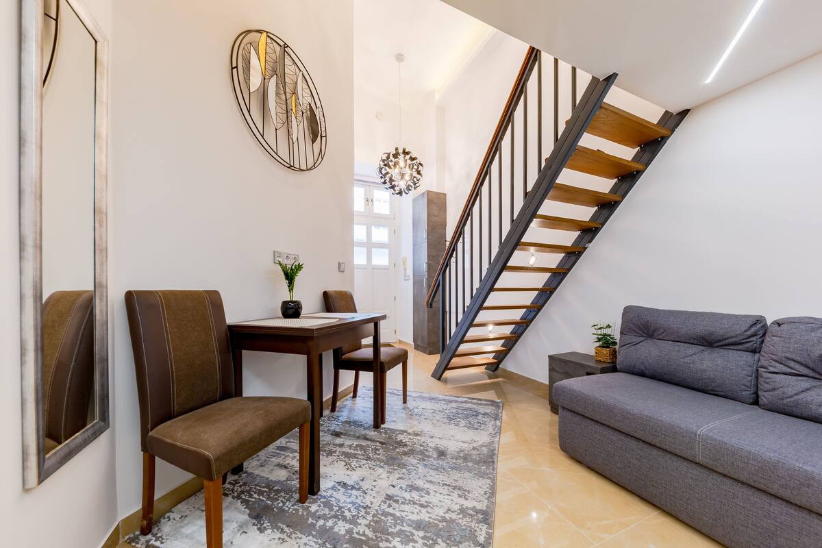 Una sala de estar moderna con un sofá gris, una mesa de comedor de madera y sillas, una escalera con escalones de madera, y un adorno en la pared.