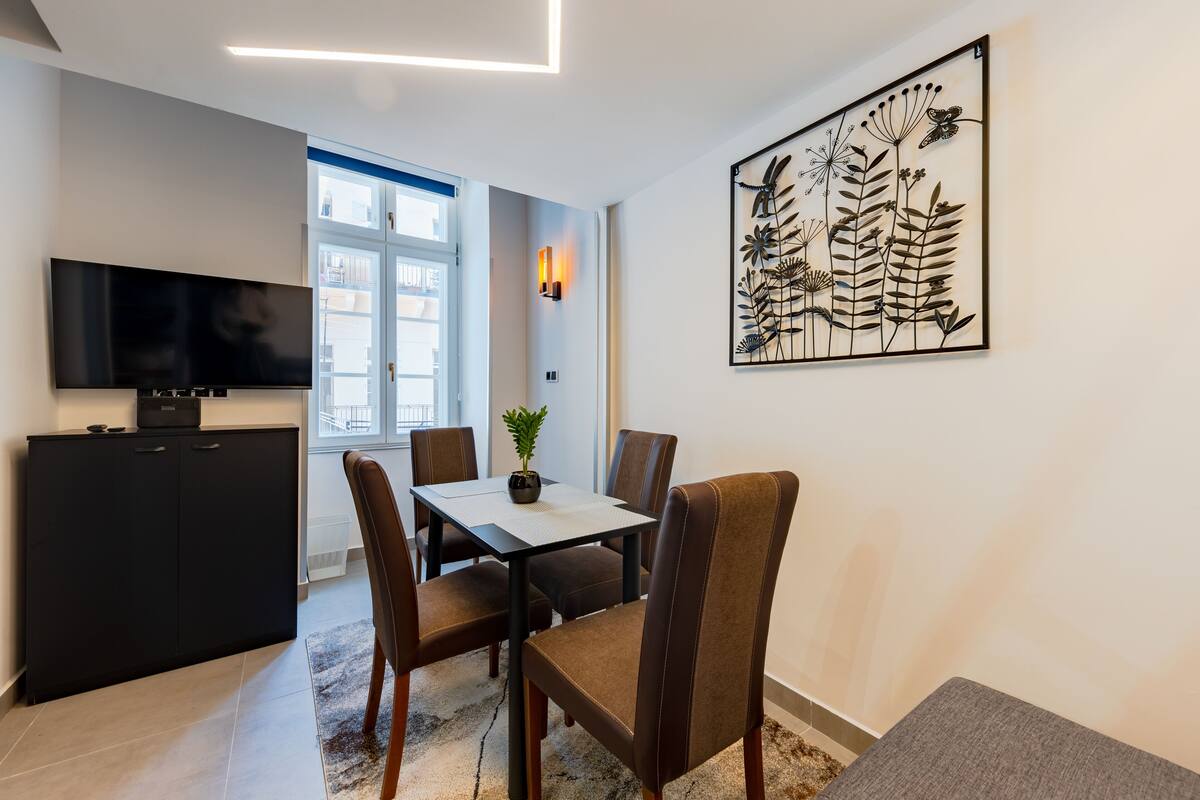 Una sala moderna con un televisor de pantalla plana, una mesa de comedor con sillas, una planta en maceta y una gran obra de arte en la pared.