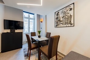 Uma sala de estar moderna com televisão de tela plana, uma mesa de jantar com cadeiras, uma planta em vaso e uma grande obra de arte na parede.