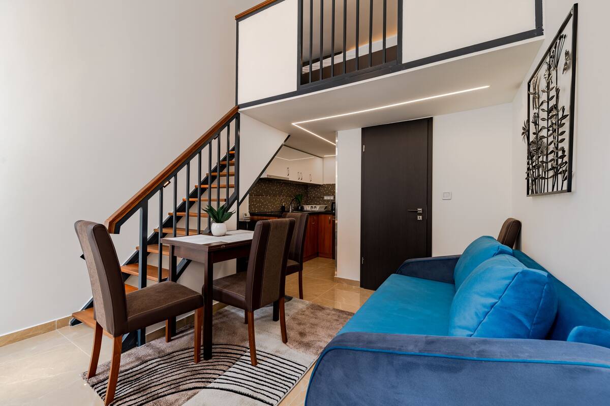 Una sala moderna con un sofá azul, una mesa de comedor de madera y sillas. Una escalera conduce a un nivel superior donde se ve una cocina.