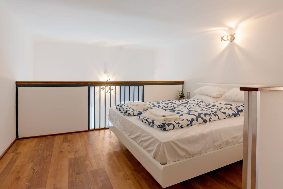 Un dormitorio con suelo de madera, una cama blanca con ropa de cama estampada y dos luces fijadas a la pared.