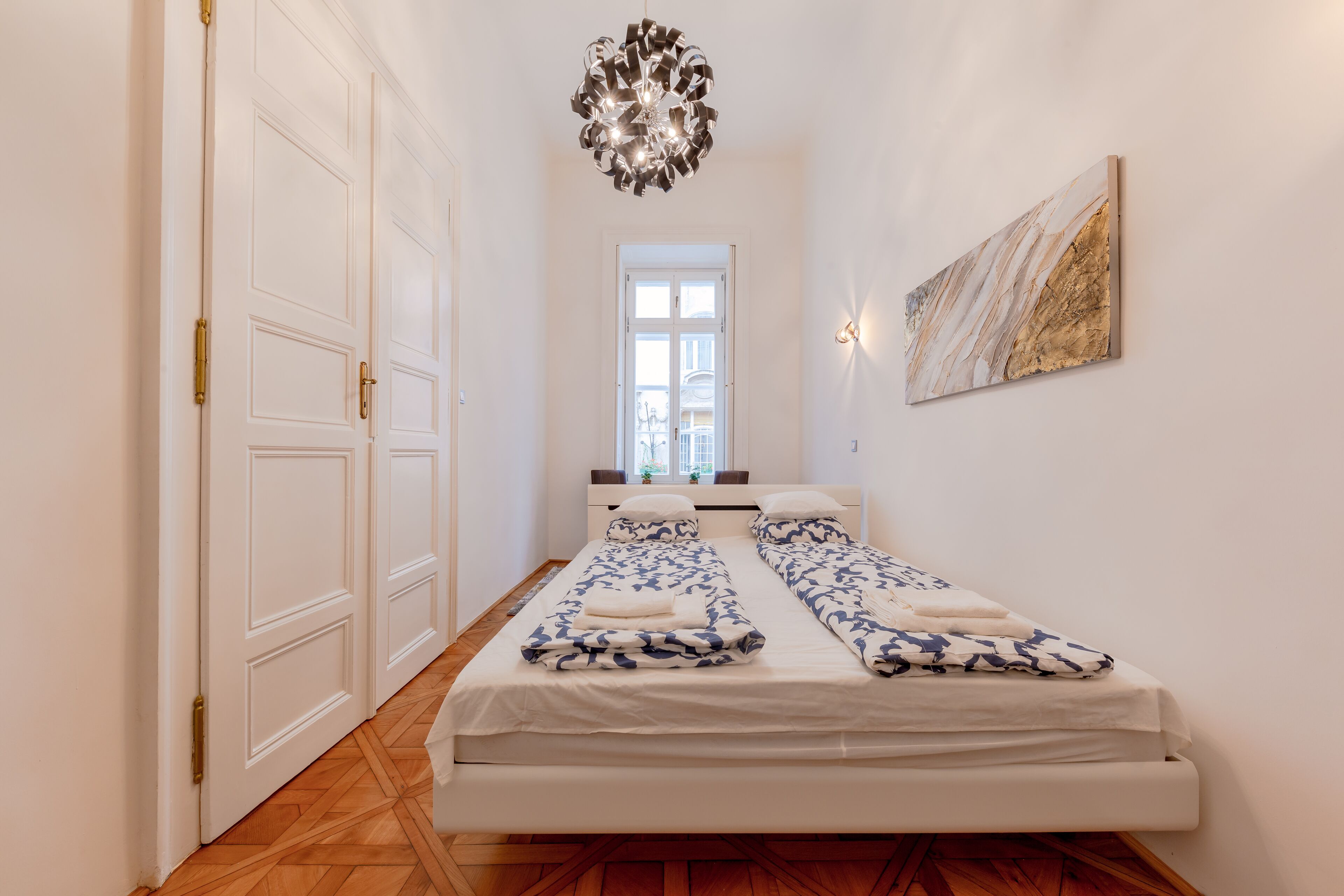 Une chambre avec deux lits, un lustre, un tableau et une vue sur l’extérieur, visible à travers une porte.