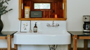 A bathroom with a white sink, a wooden-framed mirror, a framed picture, a soap dispenser, and a candle. - The Leeway - Two-Bedroom Suite (Mt Tremper)