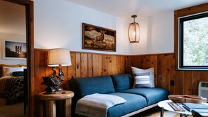 Ein gemütliches Zimmer mit einer holzverkleideten Wand, einem blauen Sofa, einer Lampe, einem Gemälde und einem Fenster mit Blick auf Bäume.