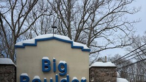Un letrero con el texto 'Big Boulder' en letras azules grandes, rodeado de terreno y árboles cubiertos de nieve.