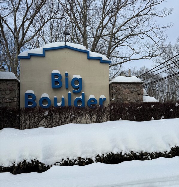 Un letrero con el texto 'Big Boulder' en letras azules grandes, rodeado de terreno y árboles cubiertos de nieve.