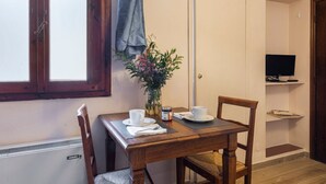 Un'area pranzo modesta con un tavolo in legno, due sedie, un vaso con fiori, una tazza bianca, un vasetto di marmellata e un televisore su uno scaffale.