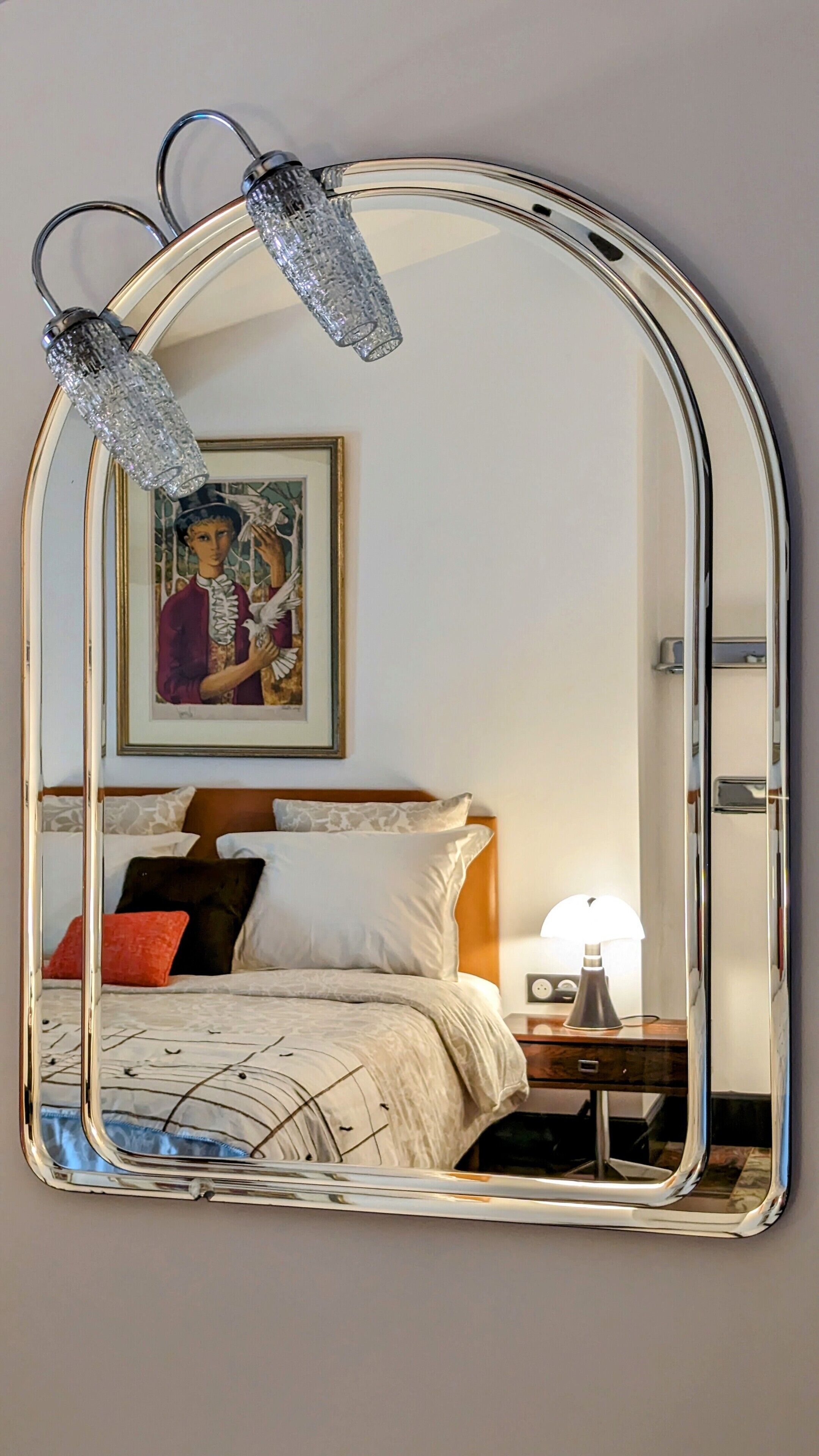 A bedroom with a large mirror, a bed with patterned bedding, a nightstand with a lamp, and a framed picture on the wall.