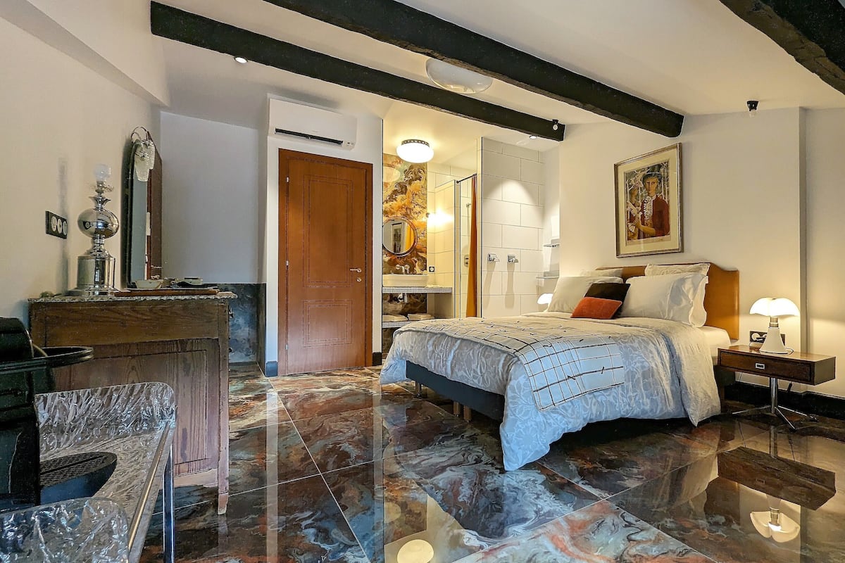 A bedroom with a bed, bedside table, lamp, and a wooden chair. There is a door leading to another room and a mirror on the wall.