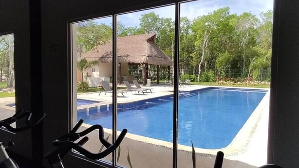 Blick aus einem Zimmer mit großem Fenster auf einen Swimmingpool, Fitnessräder und ein Gebäude mit einem Reetdach.