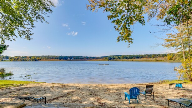 A lakeside view with a sandy beach, a row of blue Adirondack chairs, and a wooden dock extending into the water. - South Salem Vacation Rental w/ Furnished Deck! (South Salem)