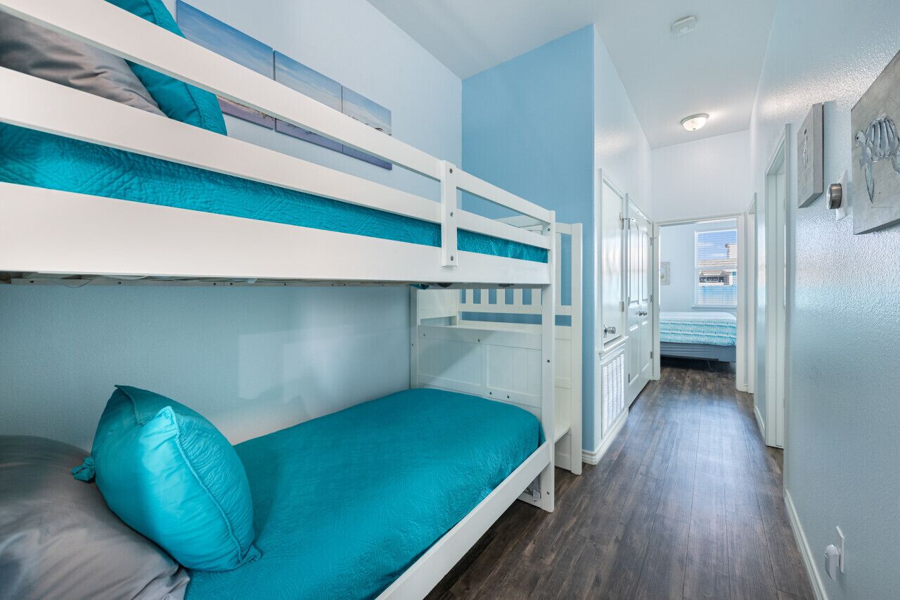 Ein Mehrbettzimmer mit türkisfarbener Matratze und Kissen, Holzboden und einem Gang zu einem weiteren Zimmer.