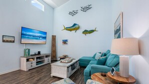 Ein Wohnzimmer mit Fernseher, einem smaragdgrünen Sofa, einem Holztisch und Wandbildern, darunter Darstellungen von Fischen und einer Strandansicht.