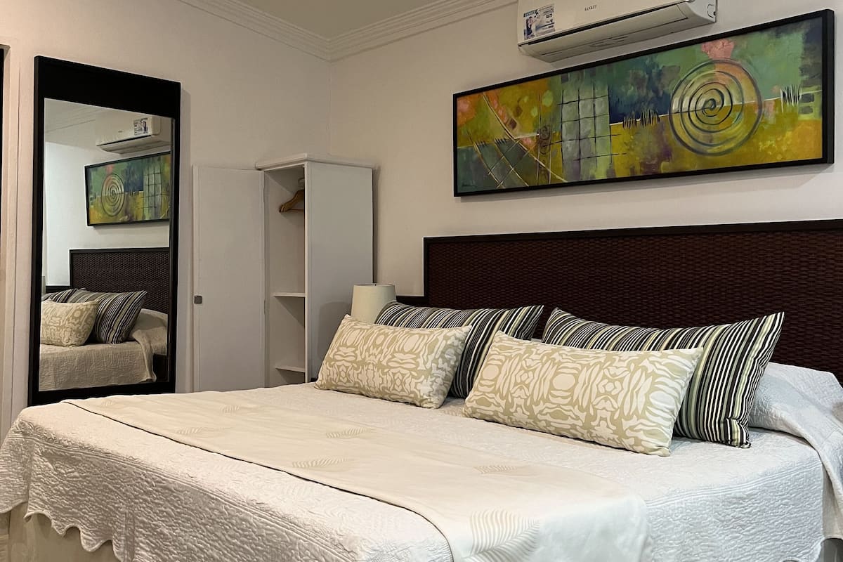 A bedroom with a large bed, a mirror, a painting, and an air conditioning unit.