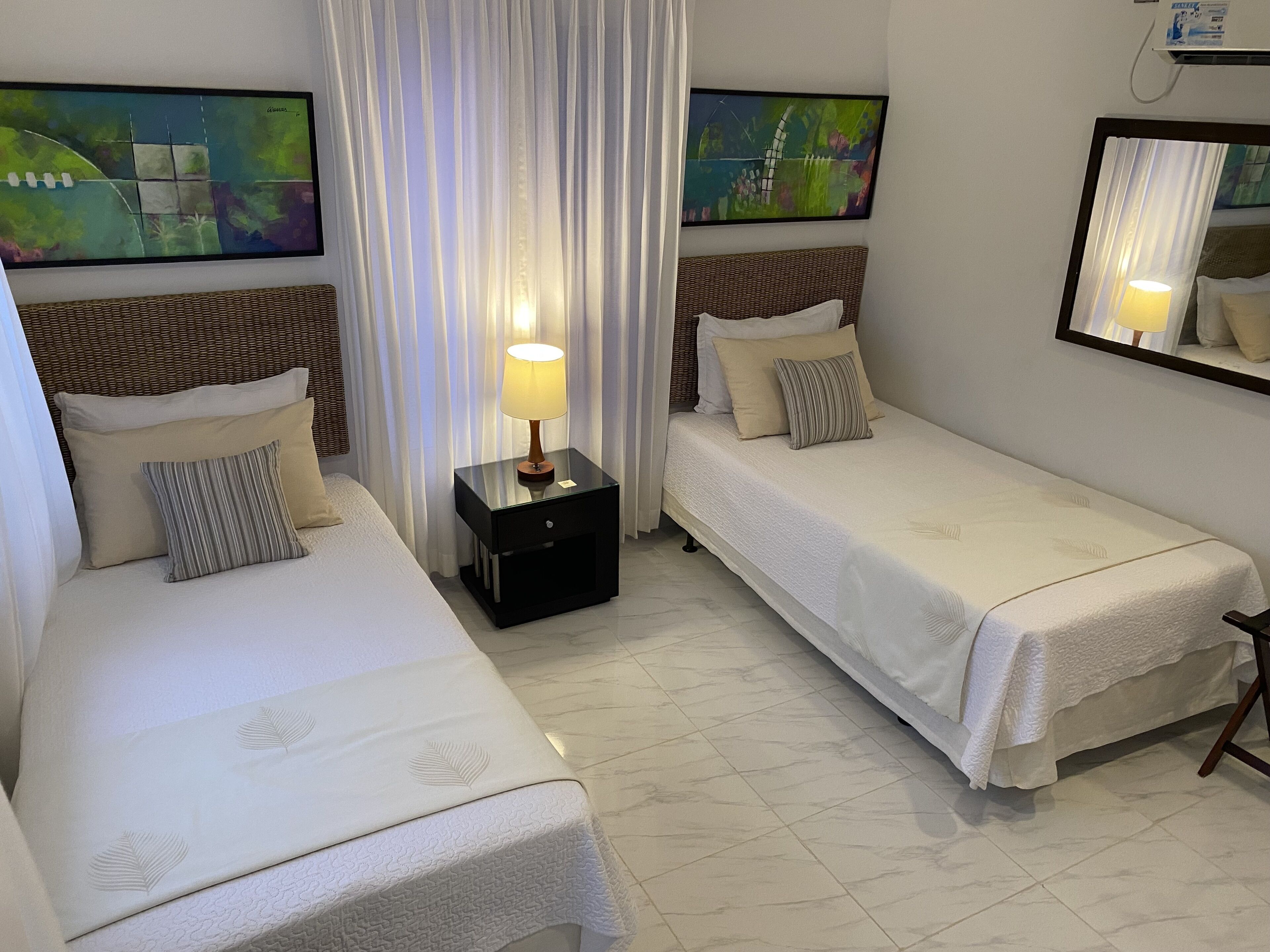 A hotel room with two single beds, a nightstand with a lamp, and two paintings on the walls.