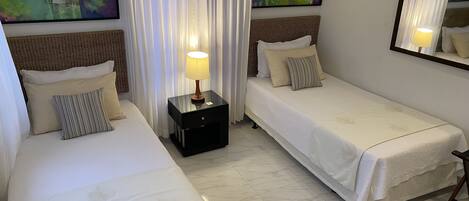 A hotel room with two single beds, a nightstand with a lamp, and two paintings on the walls.