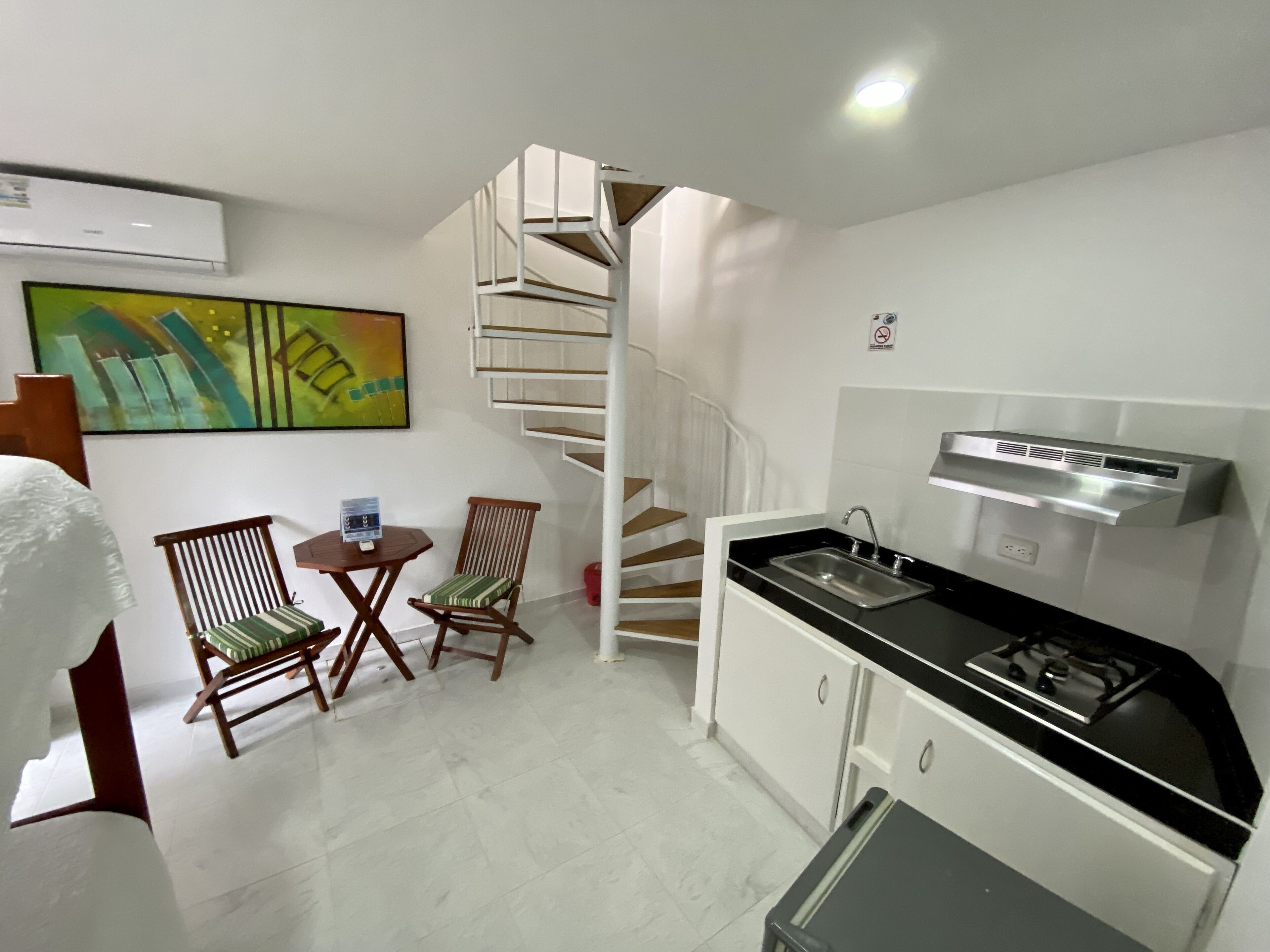 Cocina compacta con estufa a gas, un lavamanos y una pequeña zona de comedor con dos sillas y una mesa. Se observa una escalera de caracol al fondo.