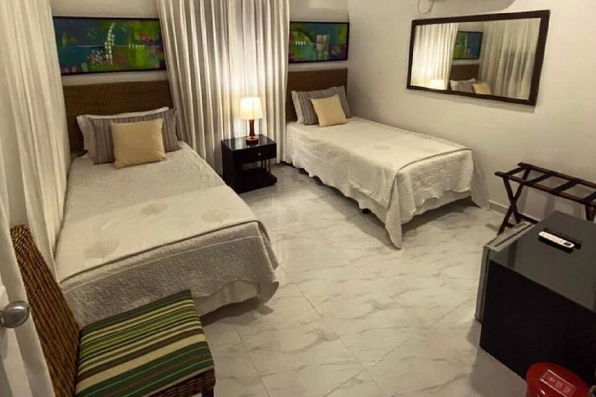 A hotel room with two beds, a nightstand with a lamp, a small table, a mirror, and an air conditioning unit.