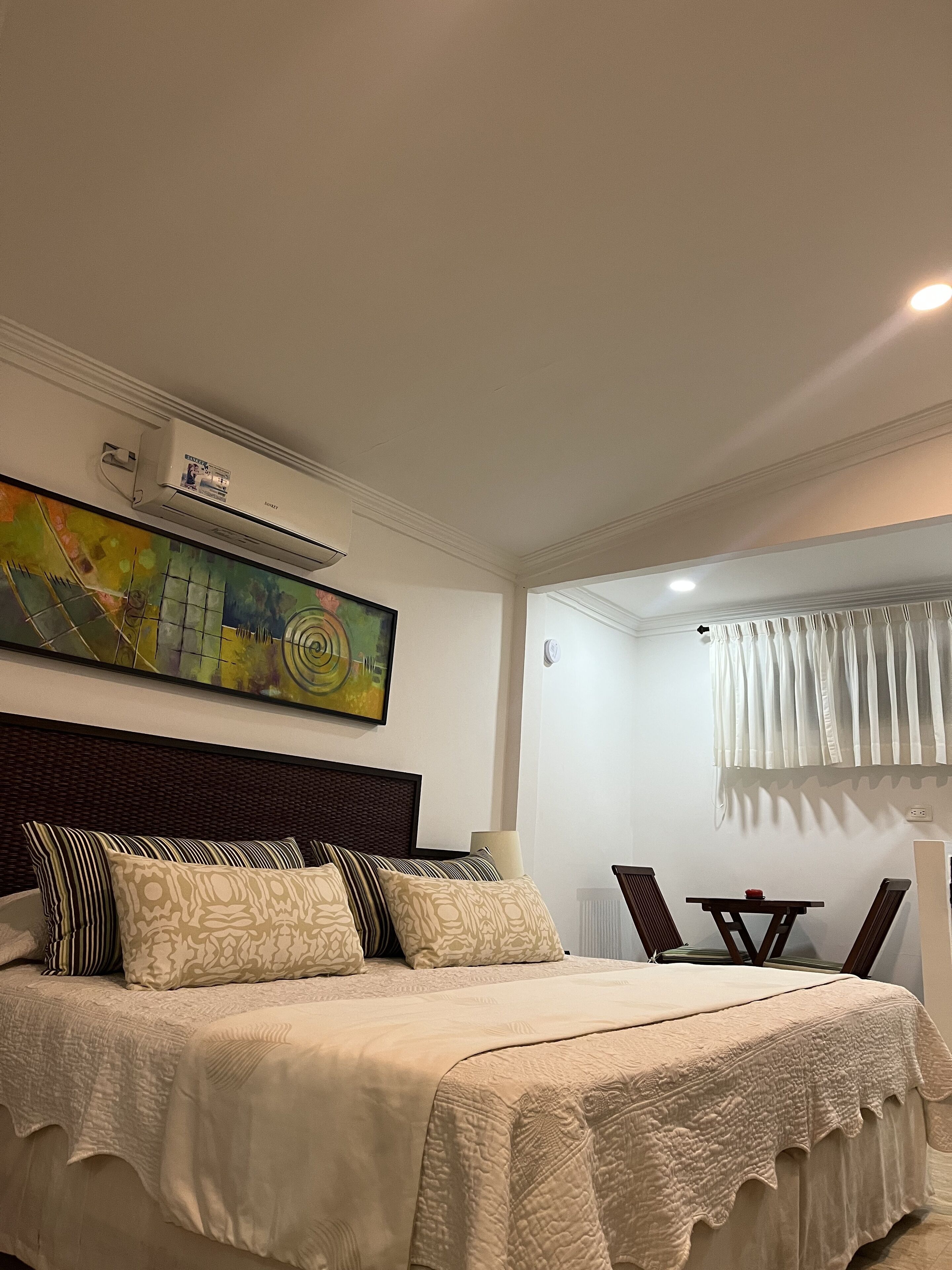 Ein Schlafzimmer mit einem großen Bett, einem Kopfteil, einer Wand-Klimaanlage, einem Gemälde und einem Fenster mit Vorhängen.