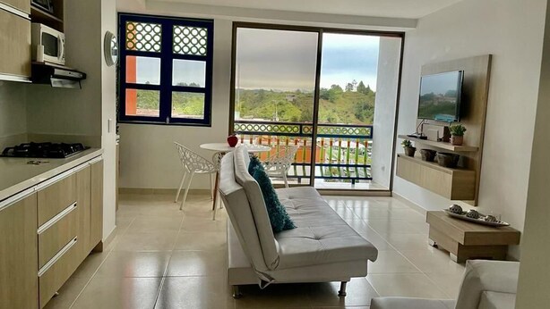 A modern living room with a sofa, a dining table, and a TV. There is a balcony with a view of greenery. - Edificio San Jose Parque Guatape (Guatapé)