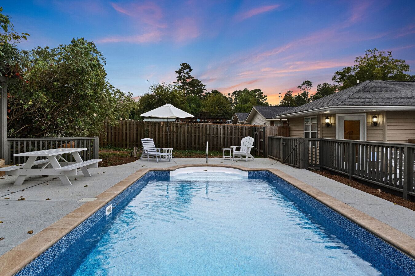 A rectangular swimming pool with a wooden deck, surrounded by a wooden fence and a wooden deck. There are two white Adirondack chairs and a picnic table.