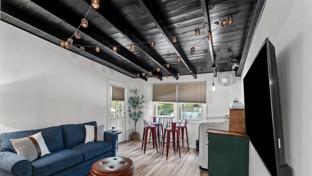 Ein modernes Wohnzimmer mit einem blauen Sofa, einem Holzesstisch mit roten Stühlen, einem großen Flachbildfernseher und einer Holzzieldecke mit sichtbaren Balken.