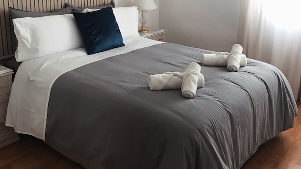 Una cama bien hecha con ropa de cama gris, una almohada blanca y una almohada azul oscuro. Hay dos toallas dobladas sobre la cama.