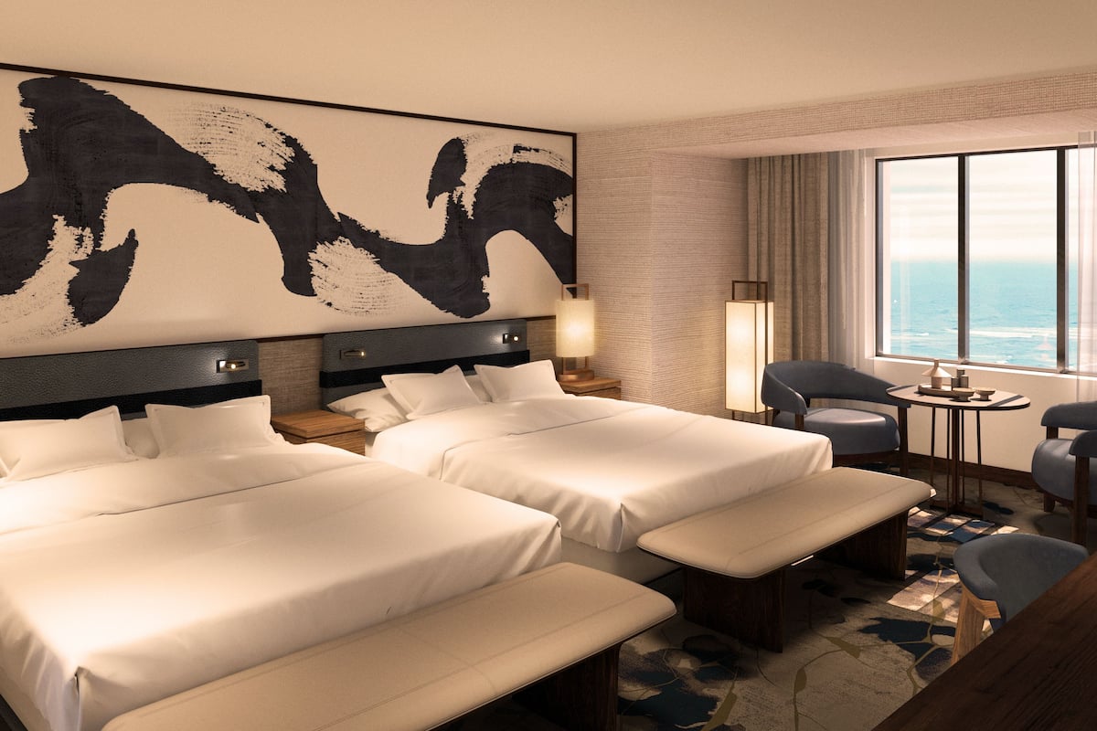 Habitación de hotel con dos camas, un cuadro abstracto de gran tamaño en la pared, vista al océano por la ventana y una pequeña mesa redonda con una lámpara.