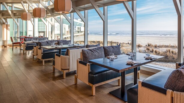 Un restaurante moderno con amplios ventanales que dan a la playa, suelo de madera y mobiliario contemporáneo.