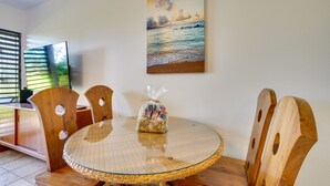 Un coin repas avec une table ronde en verre, des chaises en bois et un tableau représentant une scène de plage accroché au mur.