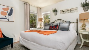 A neatly arranged bedroom with a large bed, a bedside table with a lamp, a small table with two glasses, and a chair. - Frieden Haus | 2/1, Hot Tub, Fire-pit, Full Kitchen (Fredericksburg)
