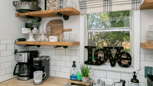 A kitchen with wooden shelves, a coffee maker, a sink, and a window with a view of trees. - Frieden Haus | 2/1, Hot Tub, Fire-pit, Full Kitchen (Fredericksburg)