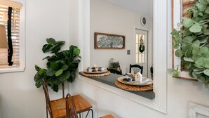 A hallway with a mirror reflecting a door, a small table with a woven tray, and large green plants. - Frieden Haus | 2/1, Hot Tub, Fire-pit, Full Kitchen (Fredericksburg)