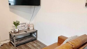 A living room with a brown leather sofa, a flat-screen TV mounted on the wall, and a small wooden side table with a plant and books.