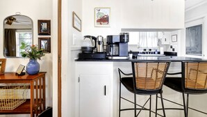 Ett modernt kök med en kaffebryggare, vattenkokare och en vas med blommor på bänkskivan. Det finns två flätade stolar och ett litet bord med en vas och en fotoram.