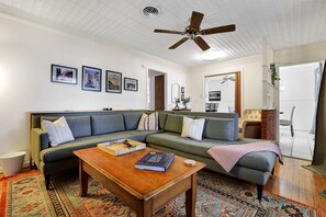 A living room with a green sofa, a wooden coffee table, and a ceiling fan. There are framed pictures on the wall and a rug on the floor.