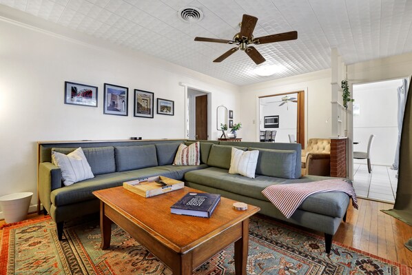 A living room with a green sofa, a wooden coffee table, and a ceiling fan. There are framed pictures on the wall and a rug on the floor.