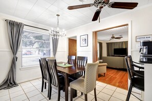 A dining area with a wooden table, chairs, and a chandelier. There is a ceiling fan, a coffee bar, and a television in the background.