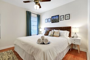 A neatly made bed with rolled towels, a wooden headboard, a bedside table with a lamp, and a ceiling fan.
